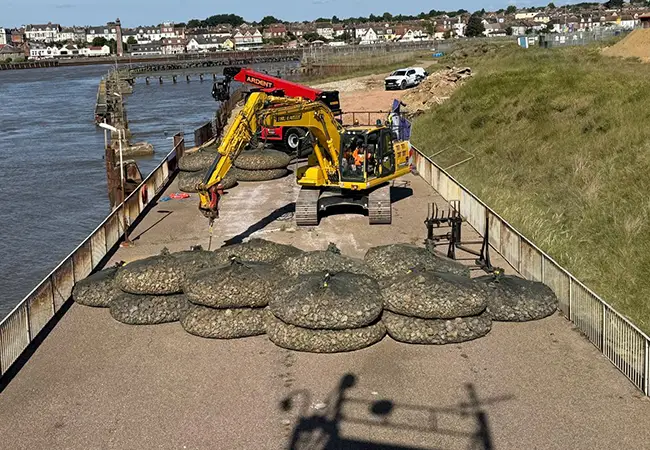 Excavator placing rock bags along a riverbank during coastal defence works